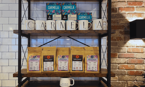 Carmela Coffee Greenwich Village display featuring bags of roasted beans, branded mugs, and merchandise arranged on wooden shelves with a brick and tile backdrop.