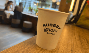 Close-up of a Hungry Ghost Coffee cup on a wooden counter inside the West Village café, with soft lighting and customers enjoying coffee in the background.