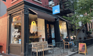 Joe Coffee West Village – The Original Since 2003 1 Exterior view of Joe Coffee in the West Village, New York City, showing the classic storefront with brown awning, outdoor bench, and customer enjoying coffee.