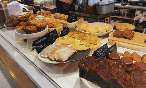 O Café Greenwich Village bakery display featuring muffins, cookies, pão de queijo, pastries, and assorted baked goods behind a glass counter.