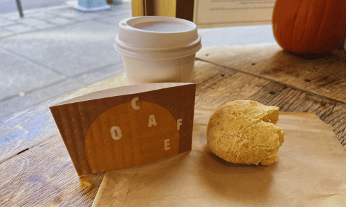Greenwich Village Coffee Shops 23 O Café Greenwich Village cup of coffee with a brown sleeve and a freshly baked pão de queijo on a wooden table by the window.