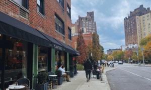 Street view of PlantShed Coffee West Village showing people enjoying coffee outdoors under black awnings, surrounded by plants and city buildings.