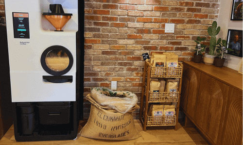 Carmela Coffee Greenwich Village small-batch roasting setup featuring a modern roaster, burlap coffee bags, retail shelves, and a brick wall backdrop.