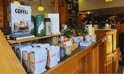 Stumptown Coffee Roasters in Greenwich Village – NYC Café 4 Stumptown Coffee Greenwich Village merch display featuring coffee bags, books, mugs, plants, and accessories arranged on a wooden counter.