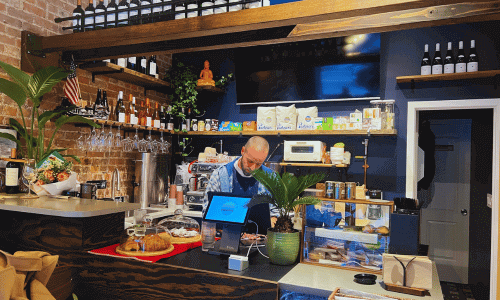 Sunriset Café West Village interior showing the barista counter with pastries, wine bottles, plants, brick walls, and a barista preparing drinks behind the register.