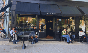 Exterior of Toby’s Estate Café West Village featuring black awnings, outdoor seating, and customers enjoying coffee along the sidewalk.