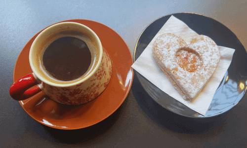 Sunriset Café West Village coffee served in a patterned mug on an orange saucer, paired with a heart-shaped powdered cookie on a black plate.