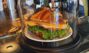 Close-up of a croissant sandwich with cheese, turkey, and fresh lettuce displayed under a glass dome at Toby’s Estate Café in the West Village.