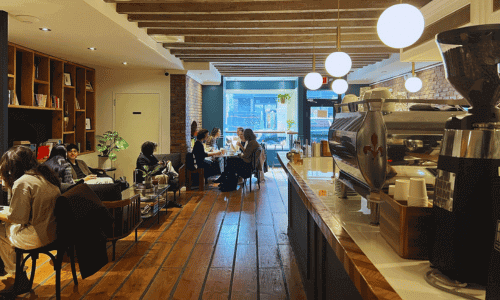 1st floor interior of Moshava Greenwich Village coffee shop with wooden floors, exposed brick walls, pendant lighting, espresso bar, and guests seated throughout the space.