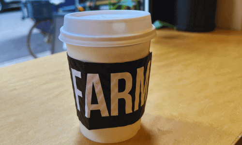 Irving Farm Greenwich Village cup of coffee with a bold black-and-white sleeve, placed on a wooden table with a street view in the background.