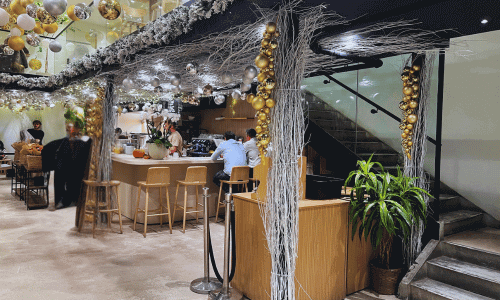 Inside Ariston Coffee Bar Greenwich Village interior featuring a modern coffee counter, hanging gold decorations, natural wood seating, and a staircase surrounded by plants.