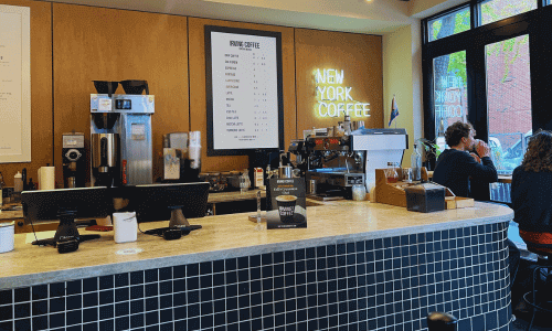 Inside Irving Farm Greenwich Village showing the marble counter, espresso machines, menu board, neon “New York Coffee” sign, and customers seated near the window.