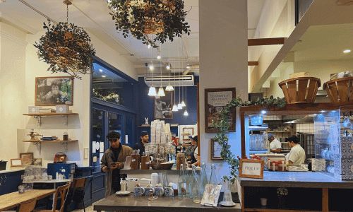 Inside Maman coffee shop Greenwich Village showing hanging greenery lights, cozy seating, wooden tables, and the busy counter area with baristas and customers.