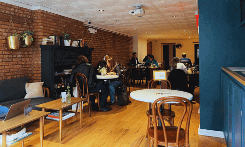 Interior 2nd floor of Moshava Greenwich Village featuring exposed brick walls, wooden floors, café tables, and guests working and relaxing in a cozy upstairs seating area.