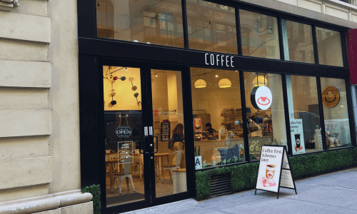 At The Venue Cafe Chelsea – Work-Friendly Coffee Shop NYC 1 Exterior view of At The Venue Cafe Chelsea with large glass windows, black framing, and sidewalk signage inviting customers.