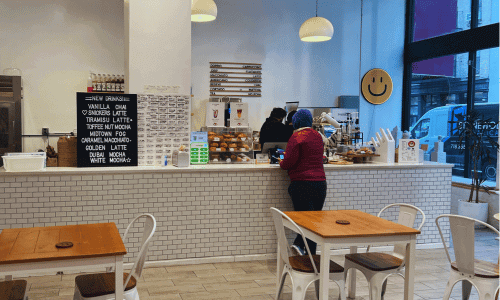 At The Venue Cafe Chelsea – Work-Friendly Coffee Shop NYC 2 Interior of The Venue Cafe Chelsea with white tile counter, pastries on display, menu board, and seating area with wooden tables.