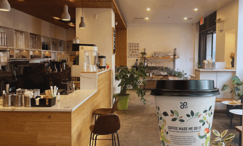 A takeaway coffee cup inside Coffee Project New York Chelsea, showing the modern wood-and-white counter area, plants, and shelves in the background.