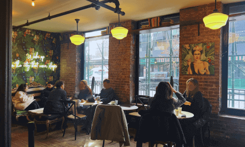 Cozy seating area inside Kobrick Coffee West Village with exposed brick walls, warm lighting, large windows, and guests enjoying coffee at small tables.