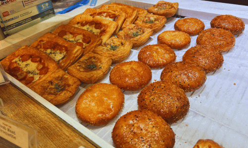 Savory pastry display at Paris Baguette in Chelsea, Manhattan, featuring baked breads, cheese pastries, and golden buns arranged on trays.