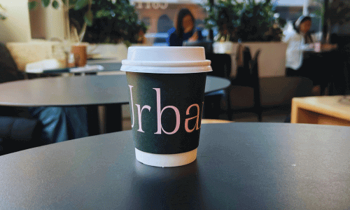 A cup of coffee on a table inside Urbana Cafe and Gallery in Chelsea, featuring a branded takeaway cup in a modern, art-filled café setting.